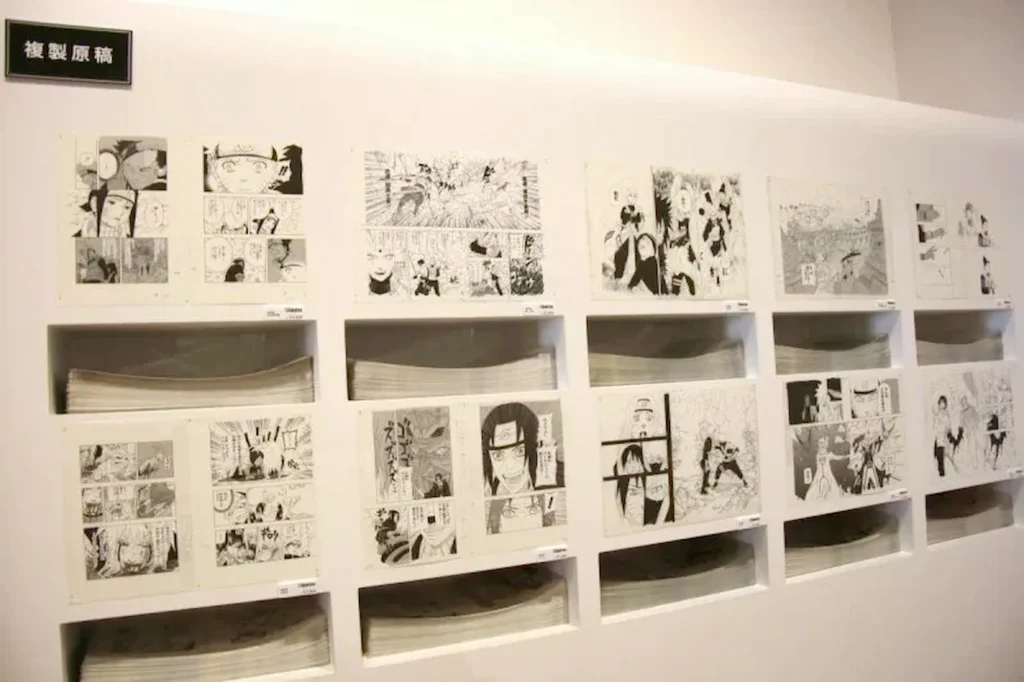 Vue d'ensemble d'une exposition officielle mettant en scène un mur de fukusei genga de Naruto par Masashi Kishimoto, avec plusieurs planches iconiques affichées au-dessus de niches lumineuses contenant d'épaisses piles de manuscrits.